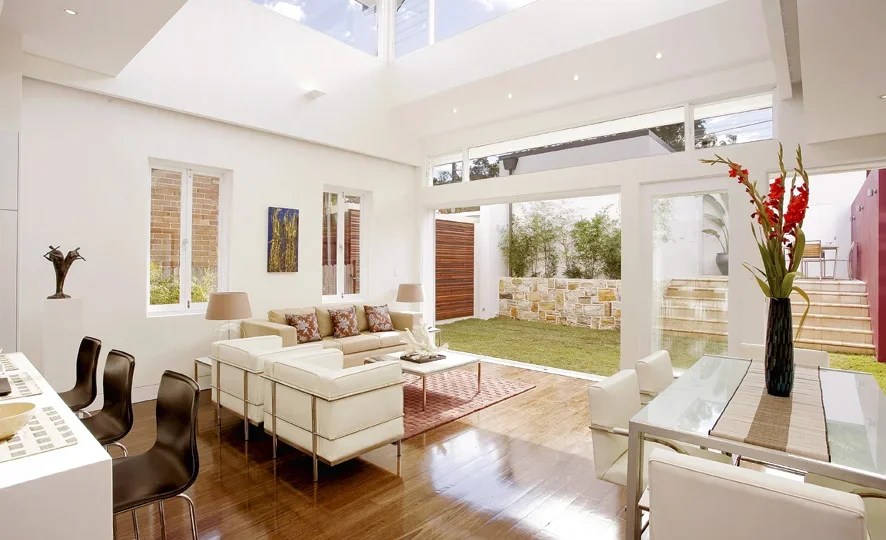 Modern living room with white walls, large windows, a beige sofa with colorful pillows, two white armchairs, a glass dining table with white chairs, and a sliding glass door opening to a backyard with grass and a stone wall.