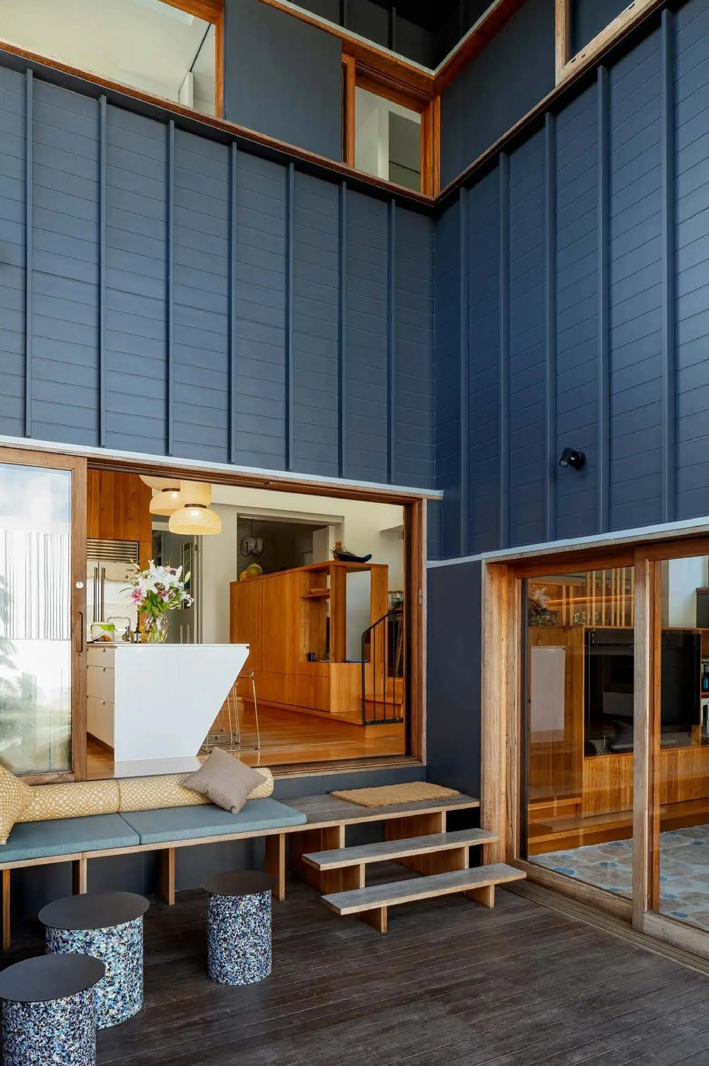Interior view of a modern house with a two-story high blue wall, wood accents, and a seating area with a bench and small tables outside a sliding glass door, showing a kitchen and staircase inside.