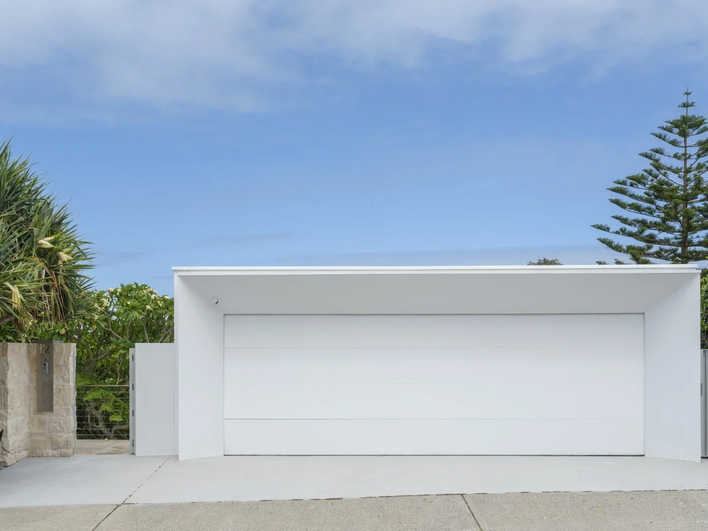 A modern white garage door with a clean, minimalist design, set against a bright blue sky with some clouds and flanked by green trees and bushes.