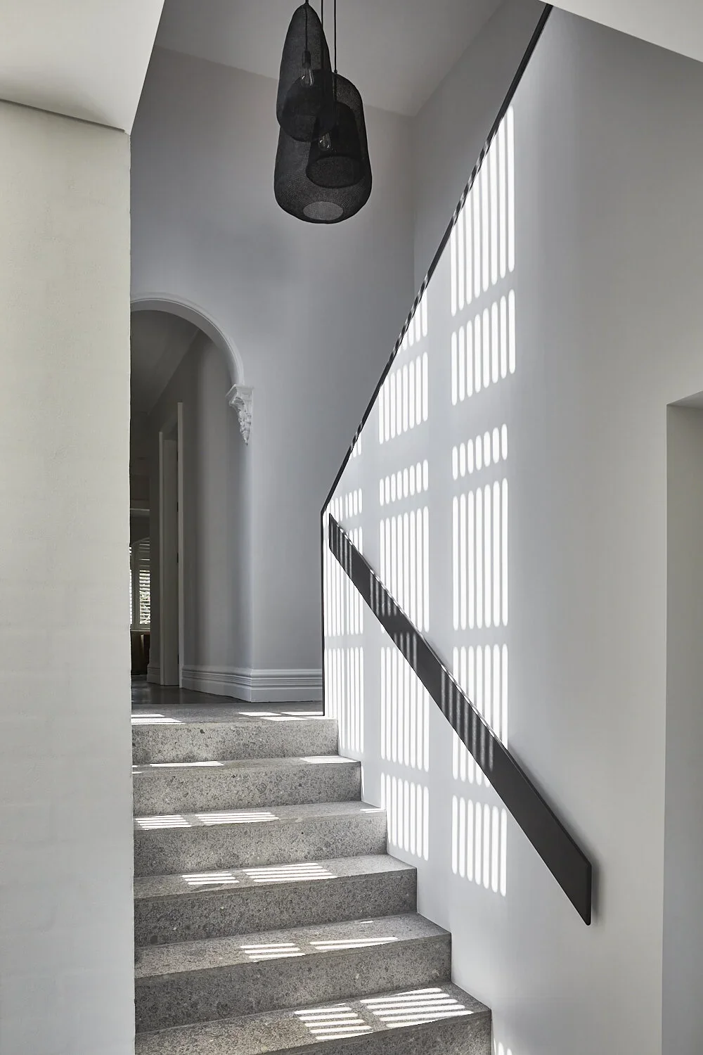A modern stairway with sunlight casting striped shadows through a window, white walls, a black handrail, and black pendant lights hanging from the ceiling.