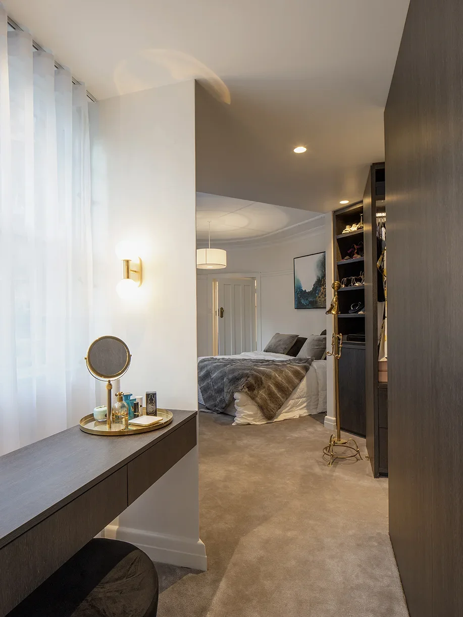 A bedroom with a bed, artwork on the wall, a dresser with toiletries, and a vanity mirror. The room has beige carpet, white and dark wood accents, and is lit with ceiling lights.
