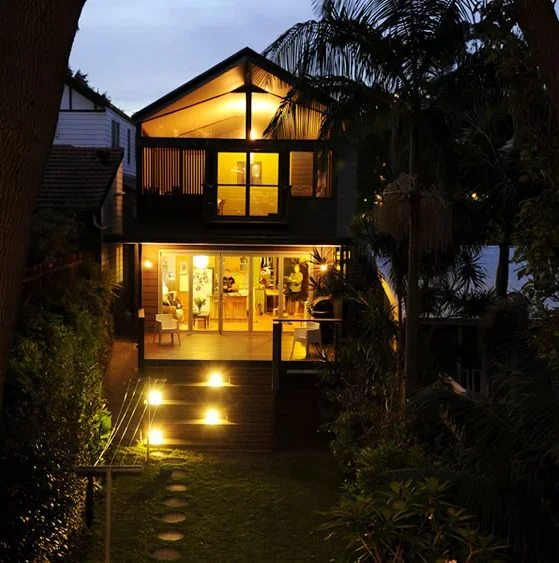 A multi-story house at night with warm interior lighting, surrounded by trees and greenery.