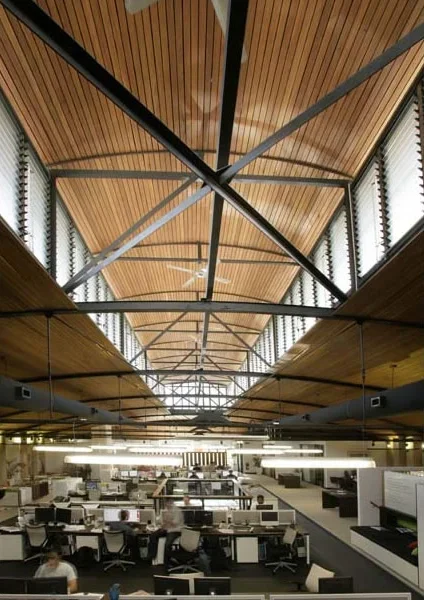 Modern office workspace with rows of desks and computers, high wooden ceiling with large windows and metal beams.