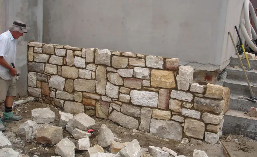 A man working on constructing a stone wall outdoors, with rocks and tools around him, near a building with concrete stairs.