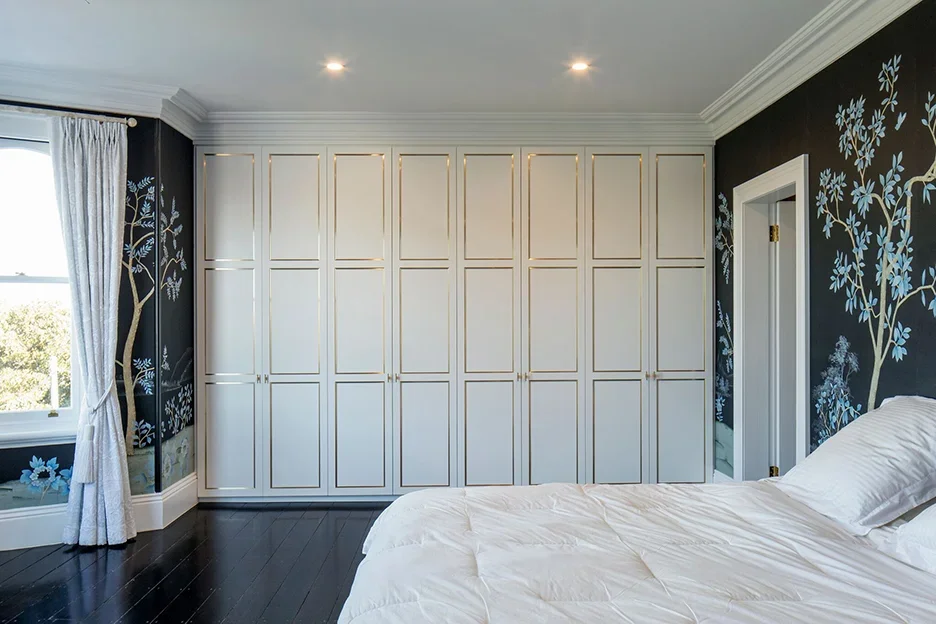 A bedroom with a white bed, a window with white curtains, black walls with blue and white floral mural, and large white wardrobe with gold hardware.