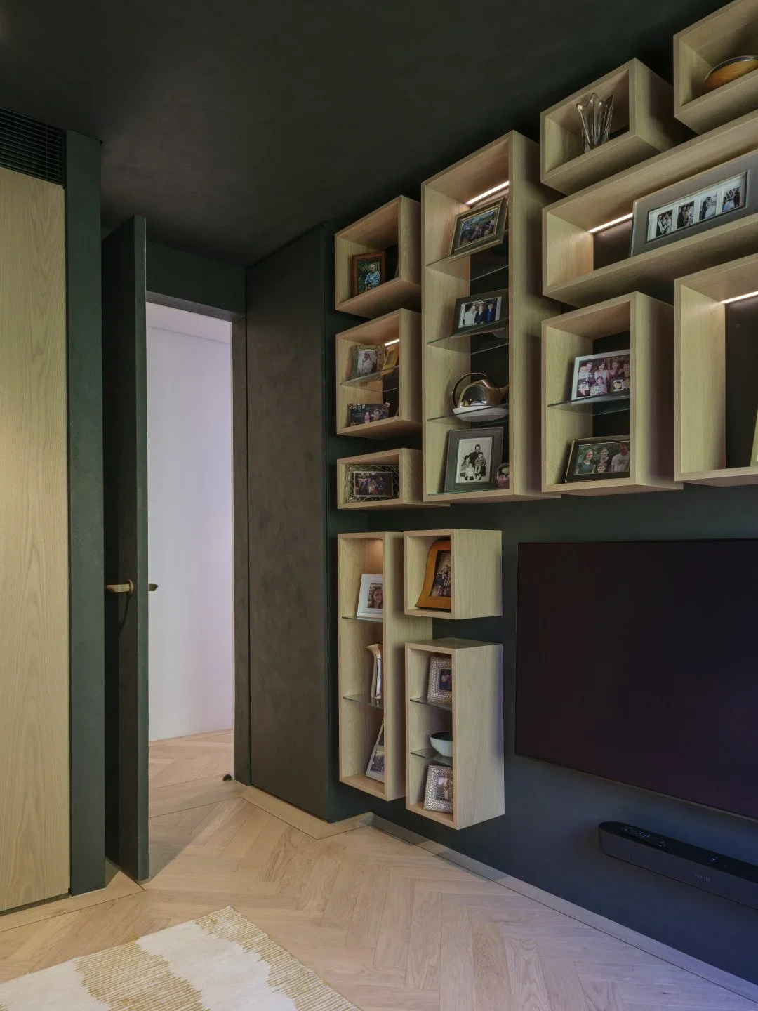Living room corner with wooden shelving unit decorated with framed photos, vases, and decorative items, dark wall, wooden floor, and TV.