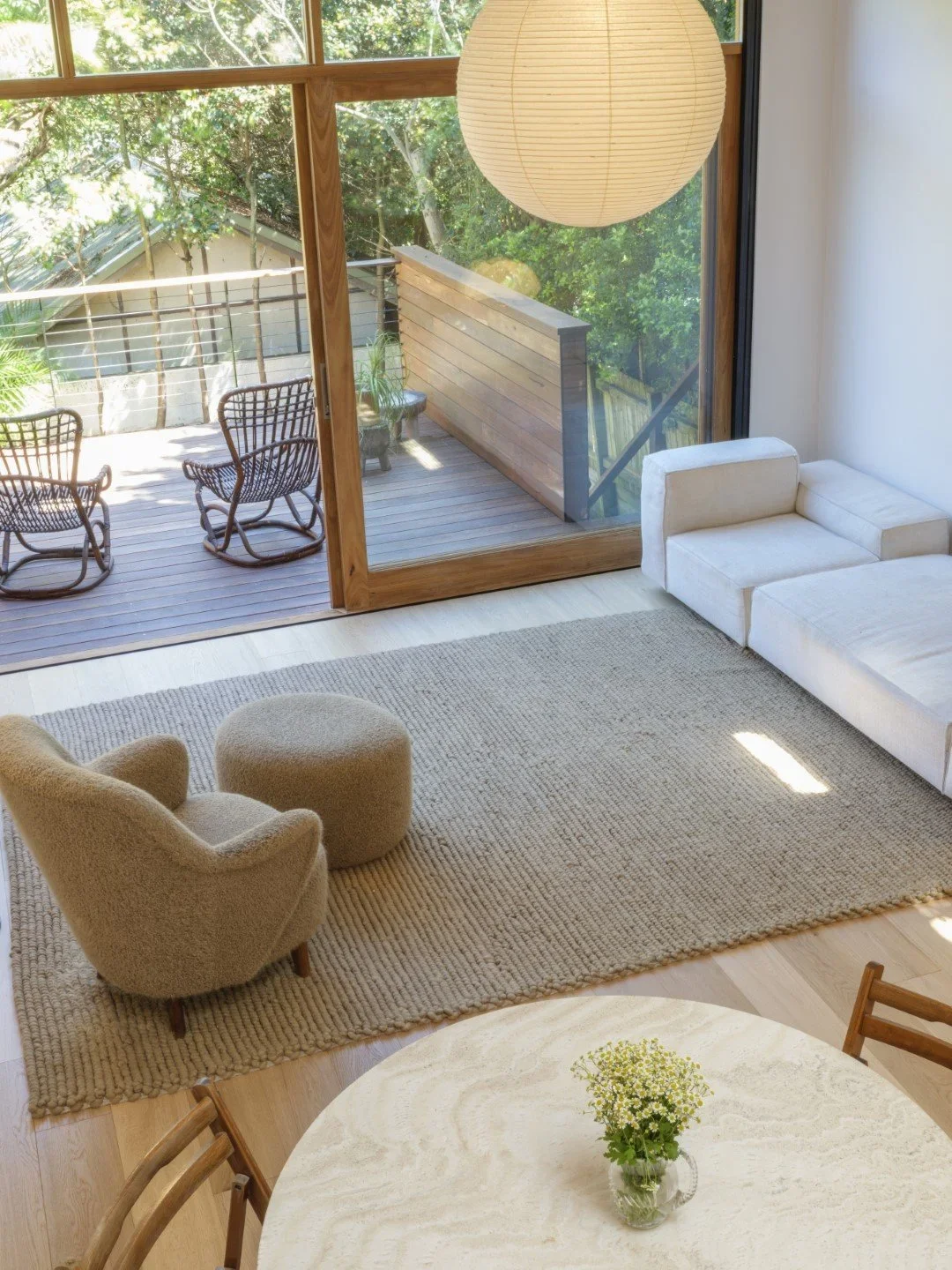Living room with a beige armchair and an ottoman on a textured rug, a white sofa, and a round dining table with a small vase of flowers. Large sliding glass door leads to a wooden deck with outdoor chairs and trees outside.
