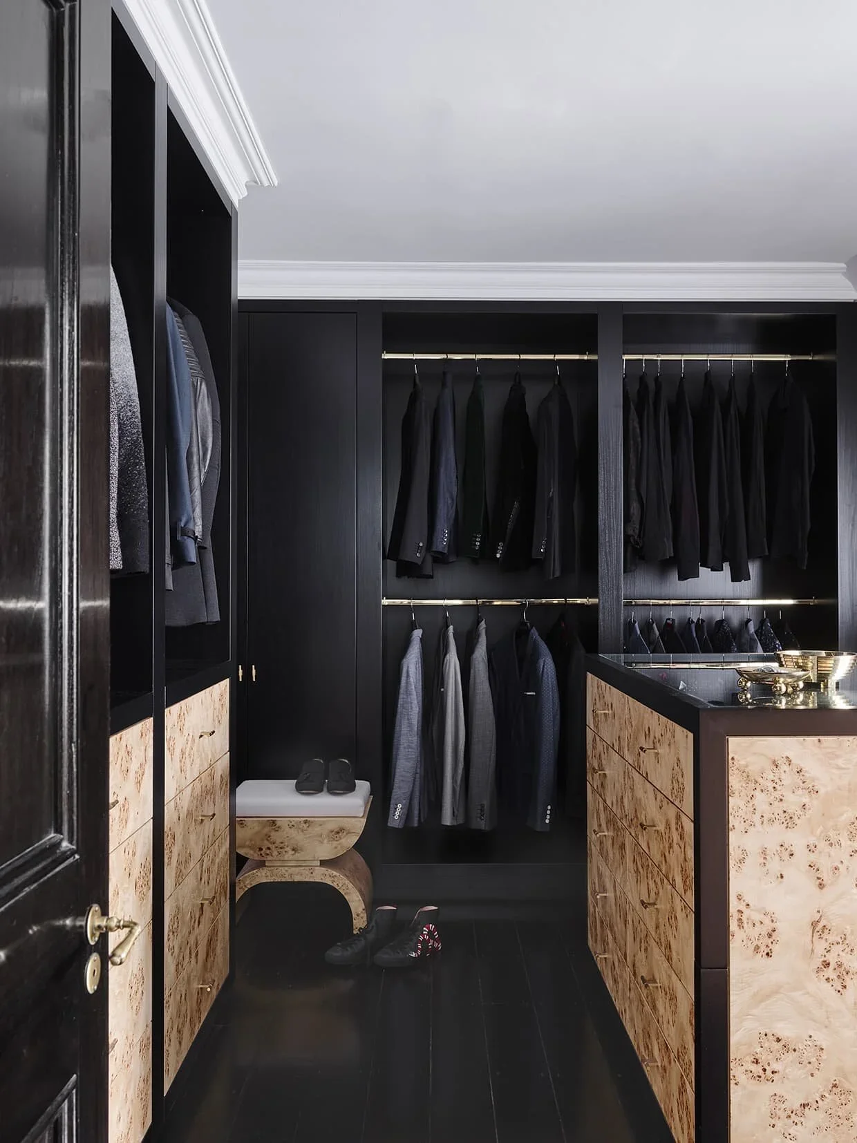 Closet with black walls and clothing racks filled with black and gray suits, with a wooden dresser and a small bench