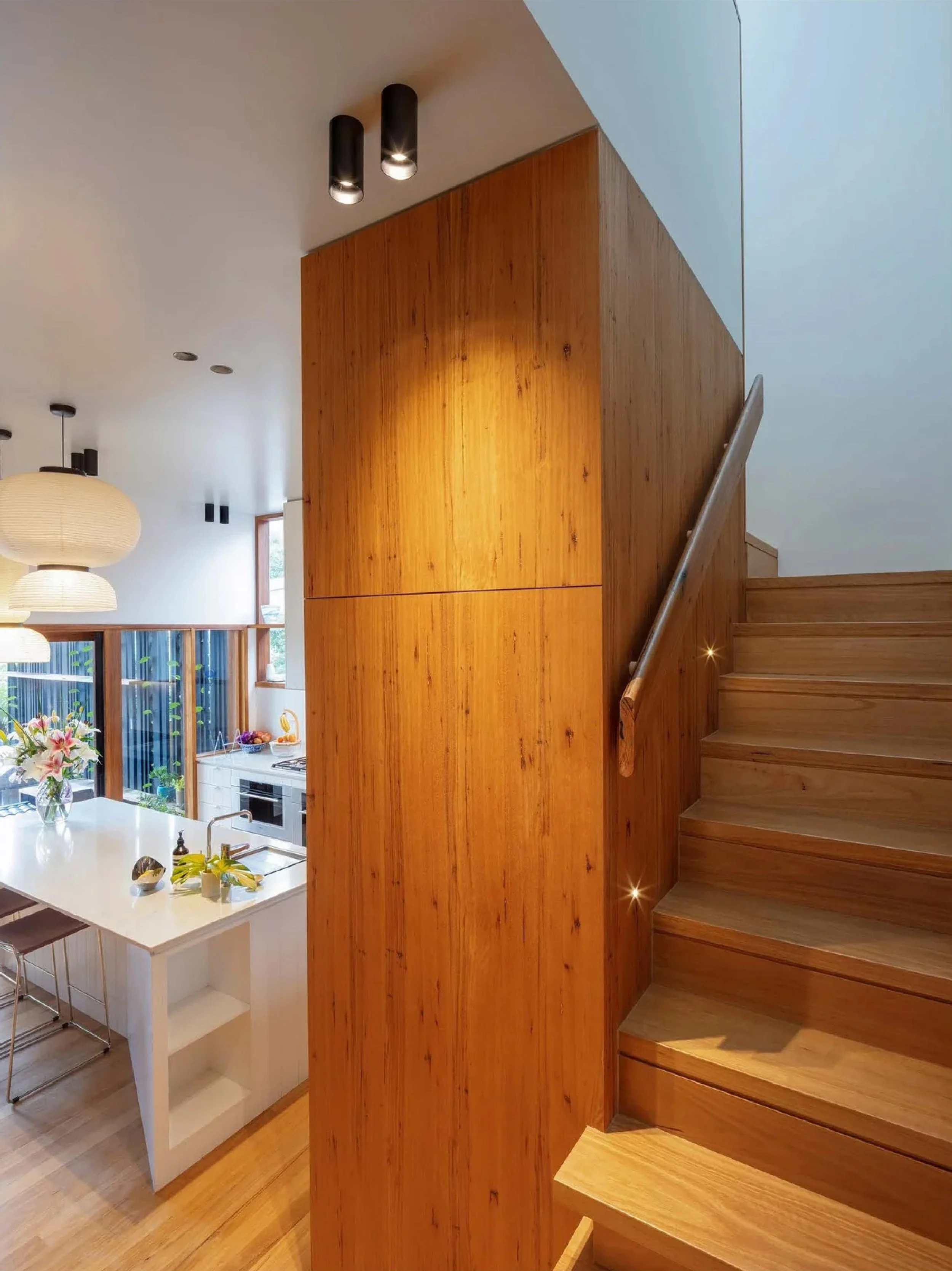 Interior view of a modern home showing a wooden staircase and wall, part of a kitchen and dining area, with hanging pendant lights and a window bringing in natural light.