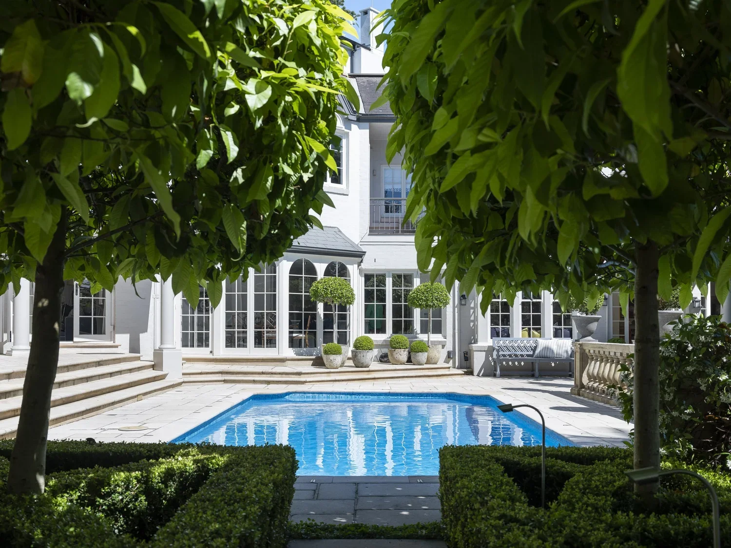 Looking through trees at a backyard with a swimming pool, surrounded by lush greenery, potted plants, and a white house with large windows and a balcony.