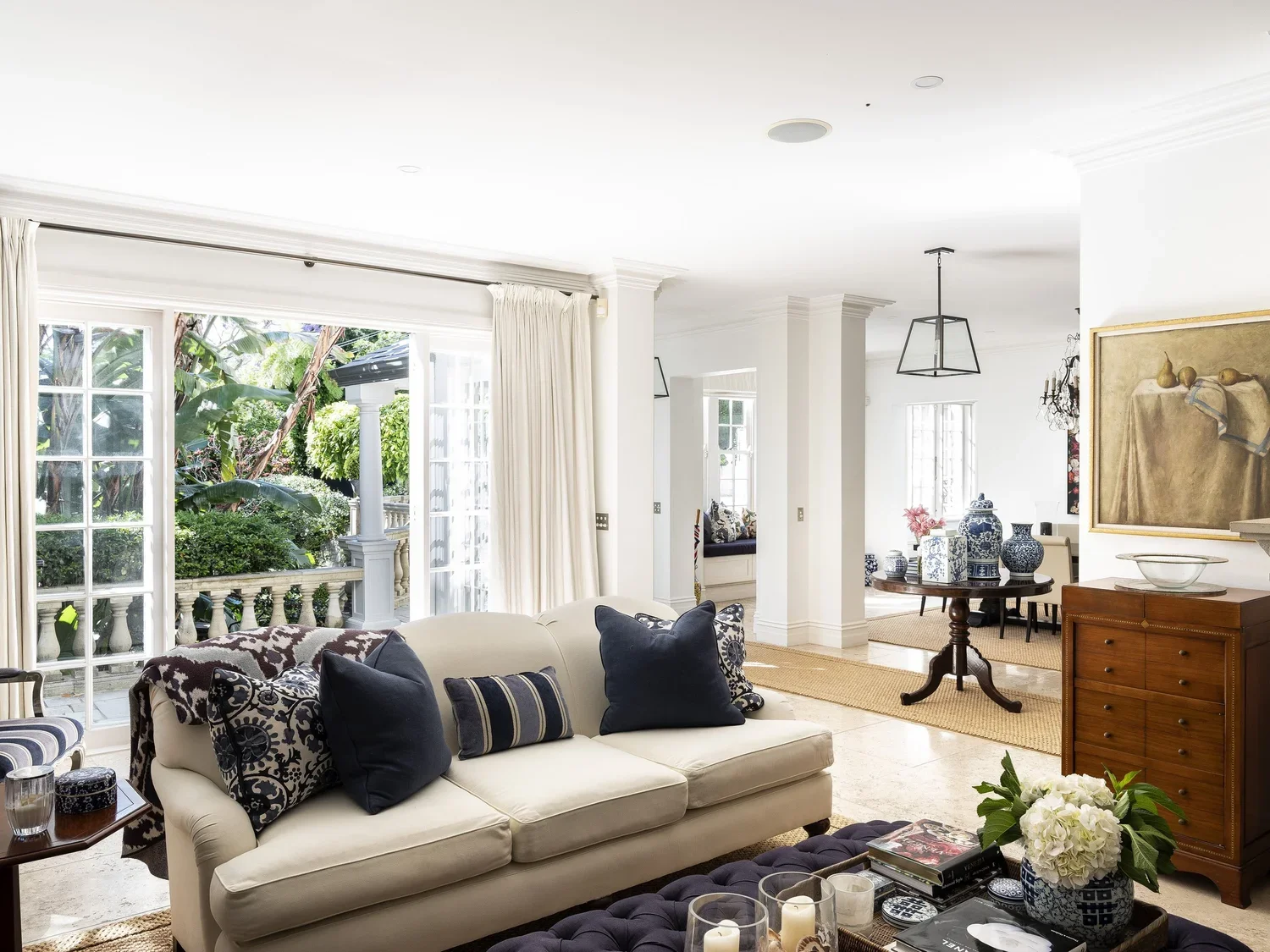 Bright living room with white walls and large windows, white sofa with blue and patterned pillows, wooden side table with candles, a wooden chest of drawers, and a round wooden table with blue and white vases, hardwood floor with beige rug, and an op