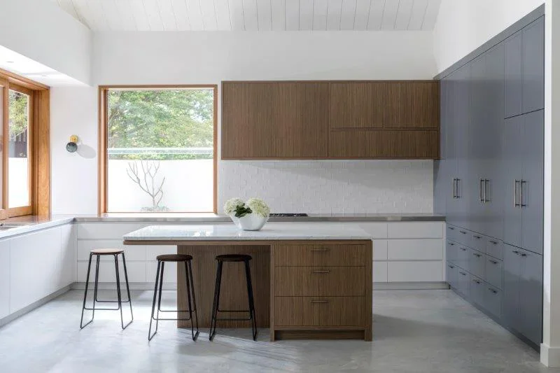 Modern kitchen with white and wooden cabinets, gray and white walls, large windows, a white countertop with a flower vase, and three black stools