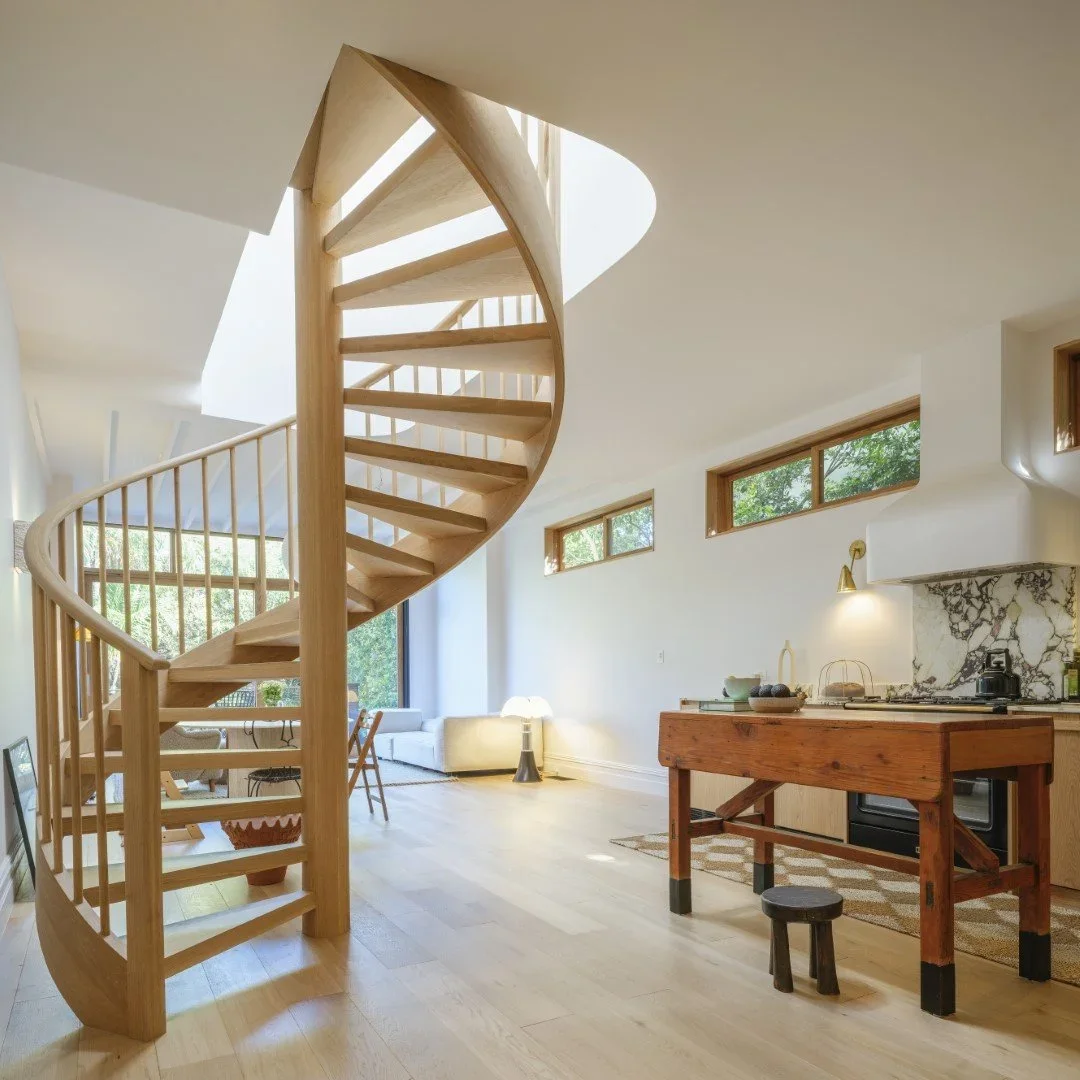 Interior view of a modern living space with a spiral staircase made of light wood, white walls, large windows, a sitting area with a white couch, and a kitchen area with wooden cabinets and a marble backsplash.