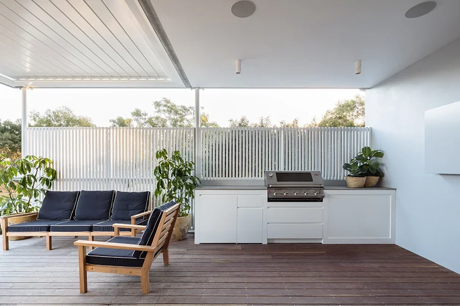 Outdoor patio with wooden flooring, a white ceiling, and a white privacy fence. Features a black outdoor sofa with cushions, a matching armchair, potted plants, and a built-in barbecue grill on the right.