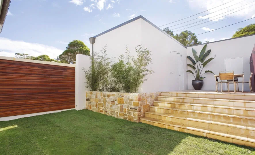 Backyard patio with yellow stone steps, white exterior wall, green plants in a planter, a wooden fence, and outdoor furniture under a partly cloudy sky.