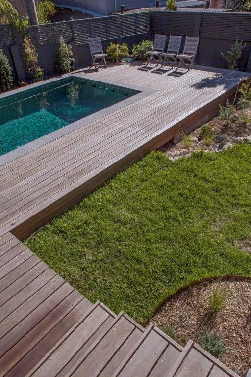 A backyard with a rectangular swimming pool surrounded by a wooden deck and a small grassy area with plants. There are four chairs on the deck near a black fence.