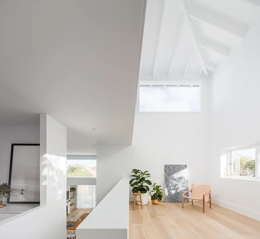 Bright living space with white walls and ceiling, large triangular window, and minimal furniture including a potted plant, a pink chair, and artwork.