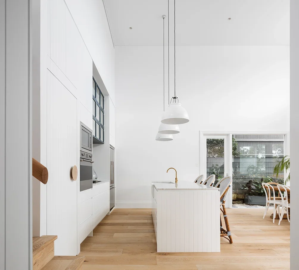Modern kitchen with white cabinets, a white island with a gold faucet, wooden bar stools, hanging white pendant lights, and large sliding glass doors leading to an outdoor garden.