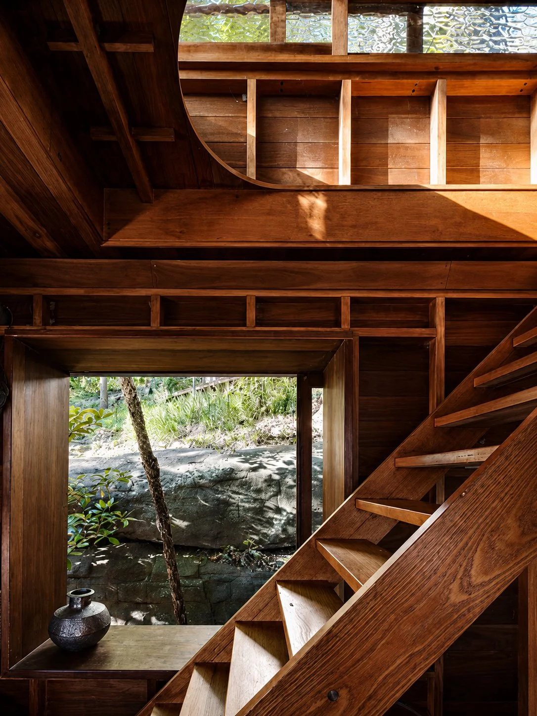 Designed by Peter Stutchbury. Interior view of a wooden cabin with a staircase leading to an upper level, large sliding glass doors opening to a lush green outdoor garden, and a wooden table with bowls on it.