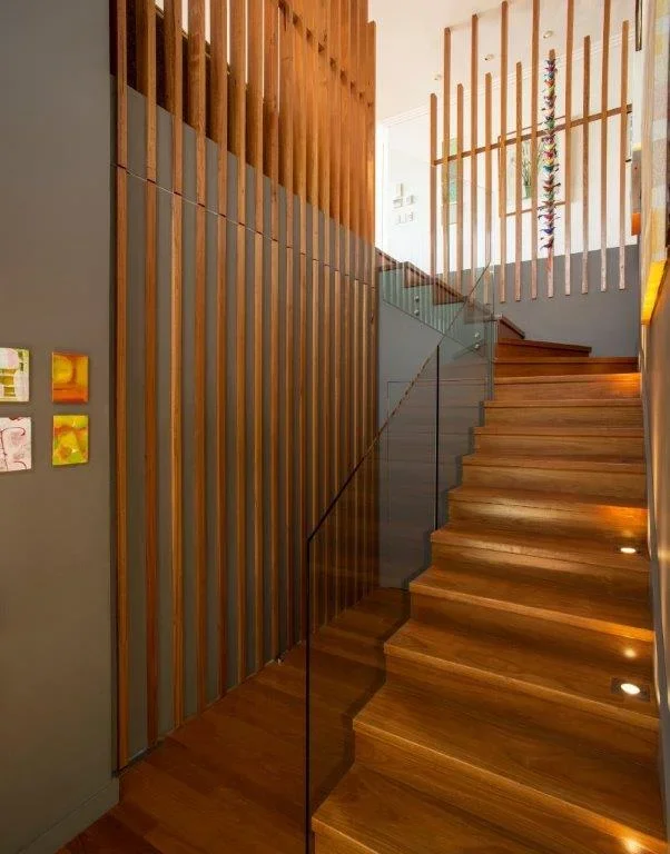 Interior staircase with wooden steps, glass railing, vertical wooden slat wall, and abstract artwork hanging on the wall in a modern home.