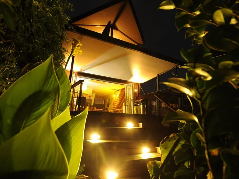 Nighttime view of a modern house with outdoor stairs leading up to an open patio, illuminated by small ground lights, surrounded by lush foliage.