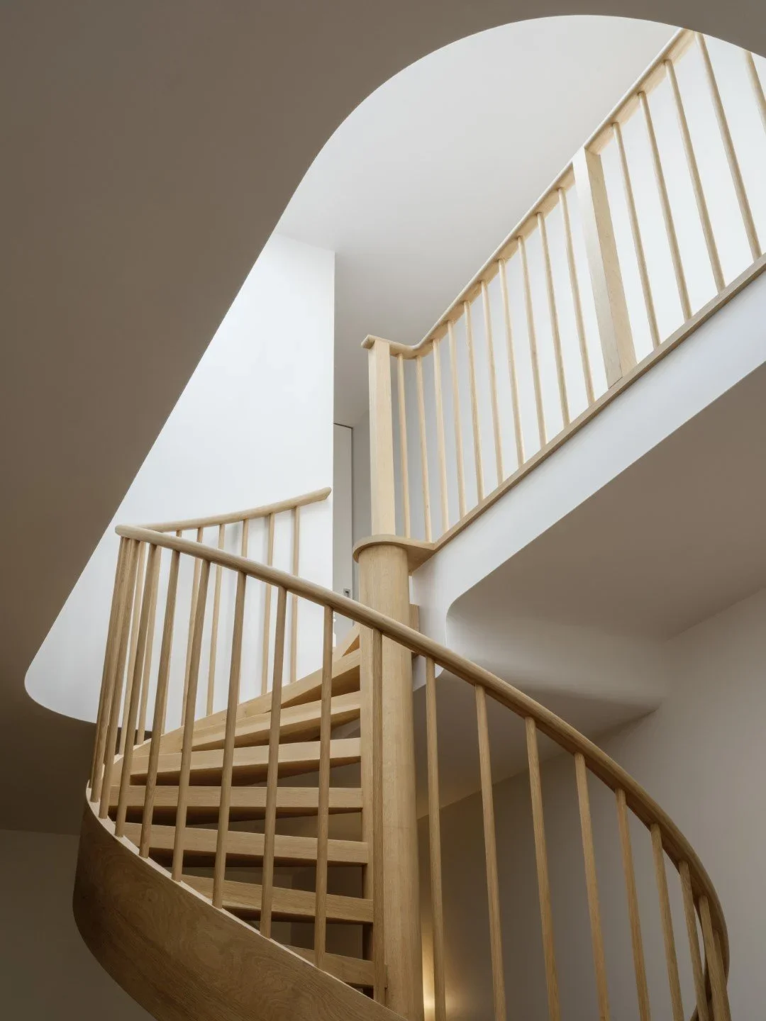 Interior view of a wooden spiral staircase with railing, seen from below, leading upward around a corner in a modern home with white walls.