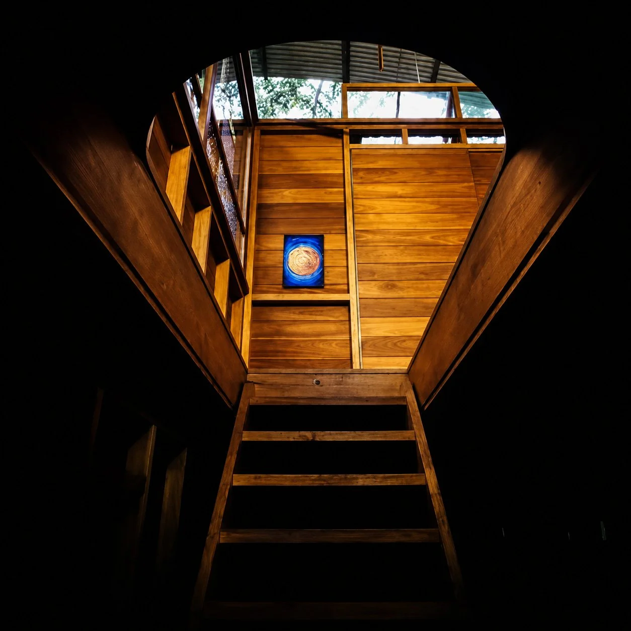 Designed by Peter Stutchbury. Interior view of a wooden cabin with a staircase leading to an upper level, large sliding glass doors opening to a lush green outdoor garden, and a wooden table with bowls on it.