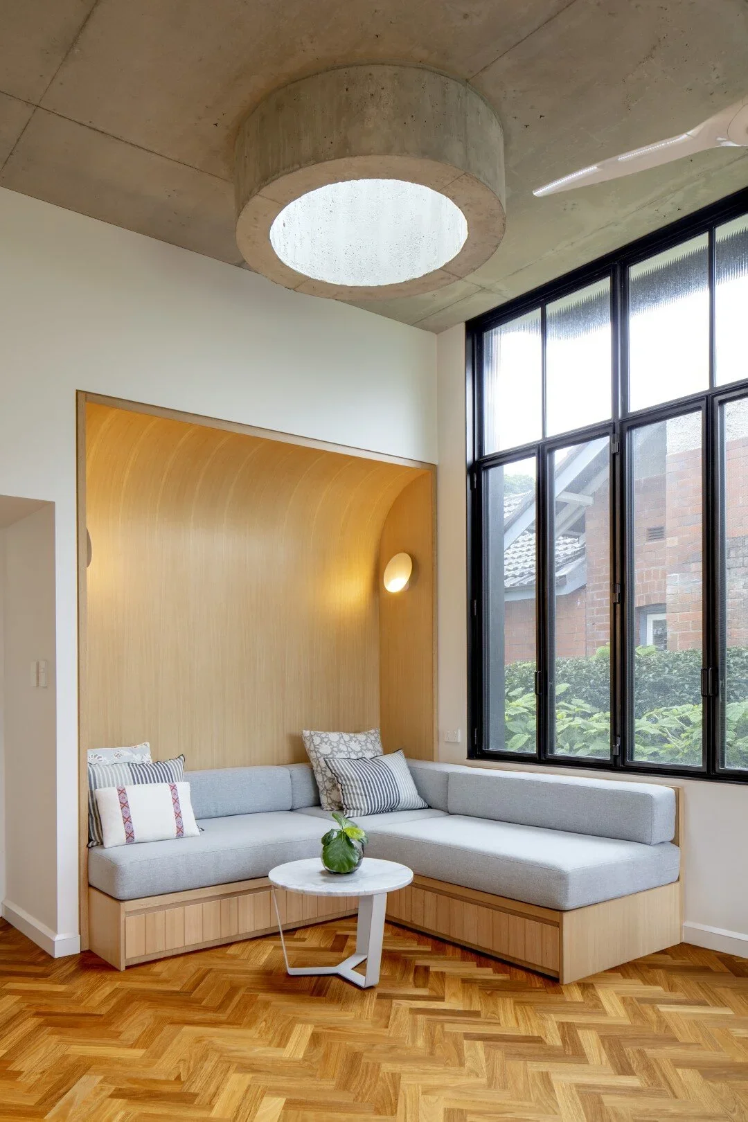Modern living room with a large corner light gray sectional sofa, patterned throw pillows, a small round white marble coffee table with a green plant, a wooden accent wall, large black-framed windows, and a concrete ceiling with a circular skylight.