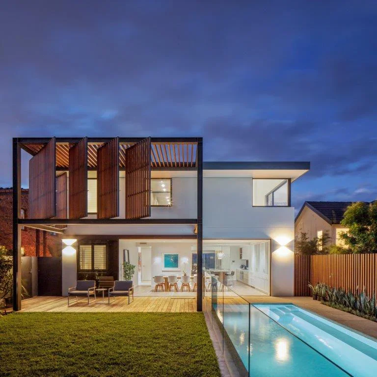 Modern two-story house with a pool in the backyard, illuminated at dusk, featuring wood and white exterior design, large glass windows, and outdoor seating area.