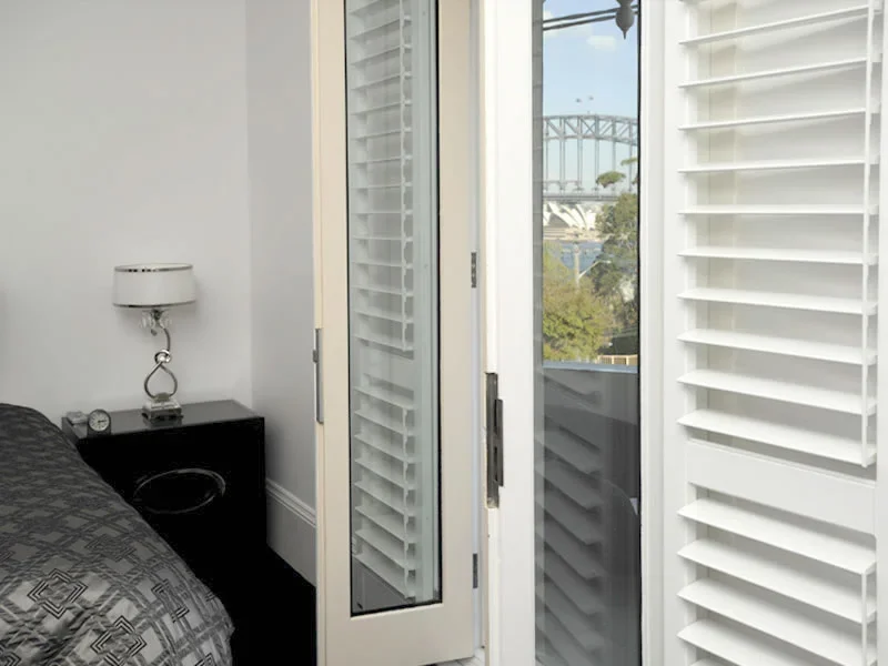 View of bedroom corner with a black nightstand and white lamp next to a bed, open balcony door showing trees and cityscape in the background.