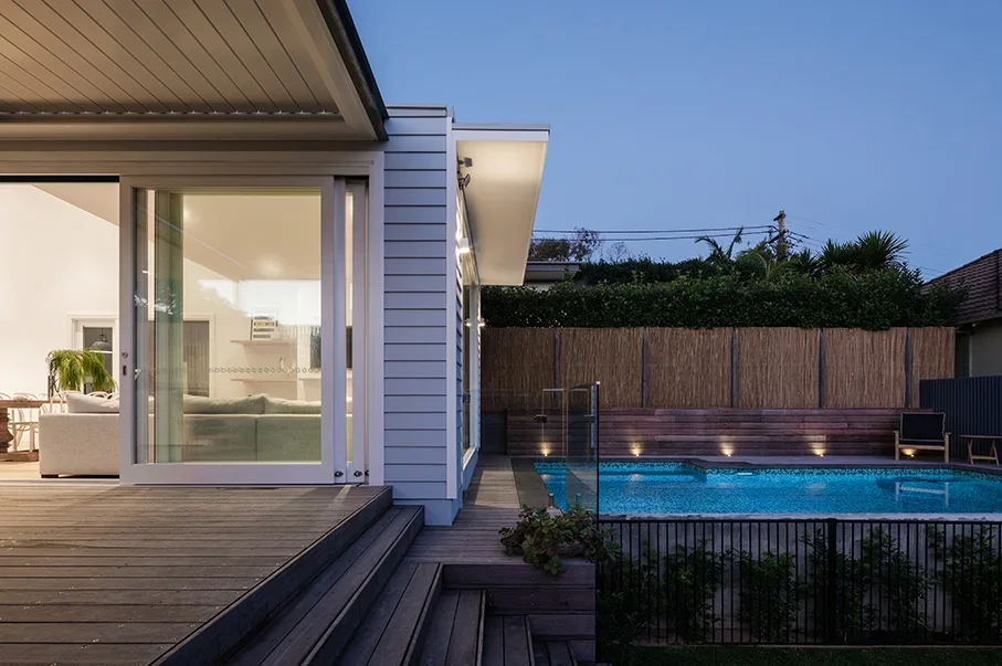 Modern house with a backyard pool at dusk, featuring a wooden deck, sliding glass door, and outdoor seating area.