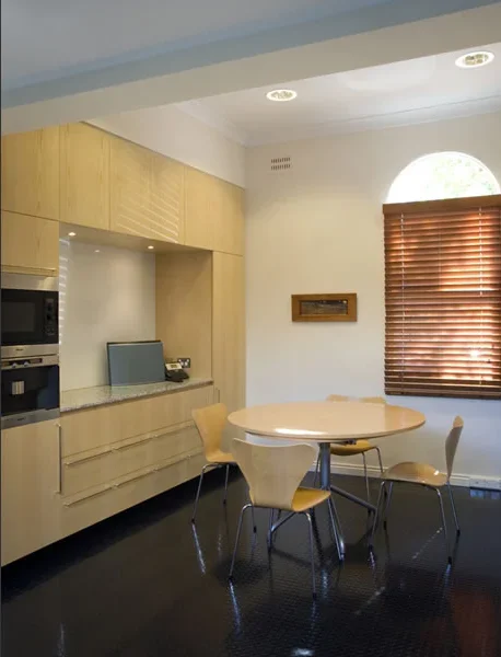 A kitchen and dining area with a round wooden table surrounded by four modern chairs. There are wooden cabinets, a microwave, and a small countertop with a phone and a few items. A window with wooden blinds is on the right, letting in natural light.