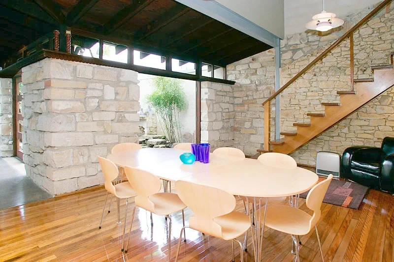 Modern dining area with a large oval white table, eight beige chairs, a stone wall, wooden staircase, and a black leather armchair.