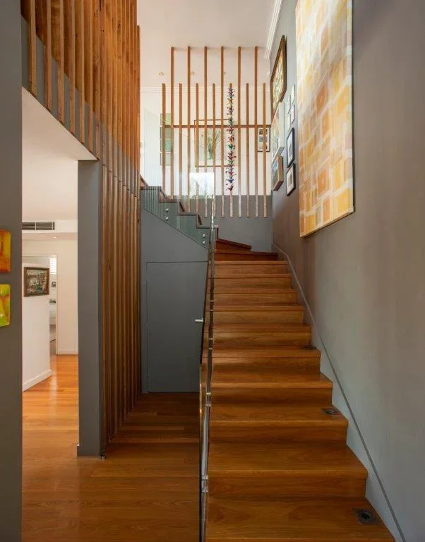 Indoor staircase with wooden steps, gray walls, and colorful artwork on the wall. A wooden railing and vertical slats create a partial divider at the top of the stairs, leading to an upper floor with large windows.