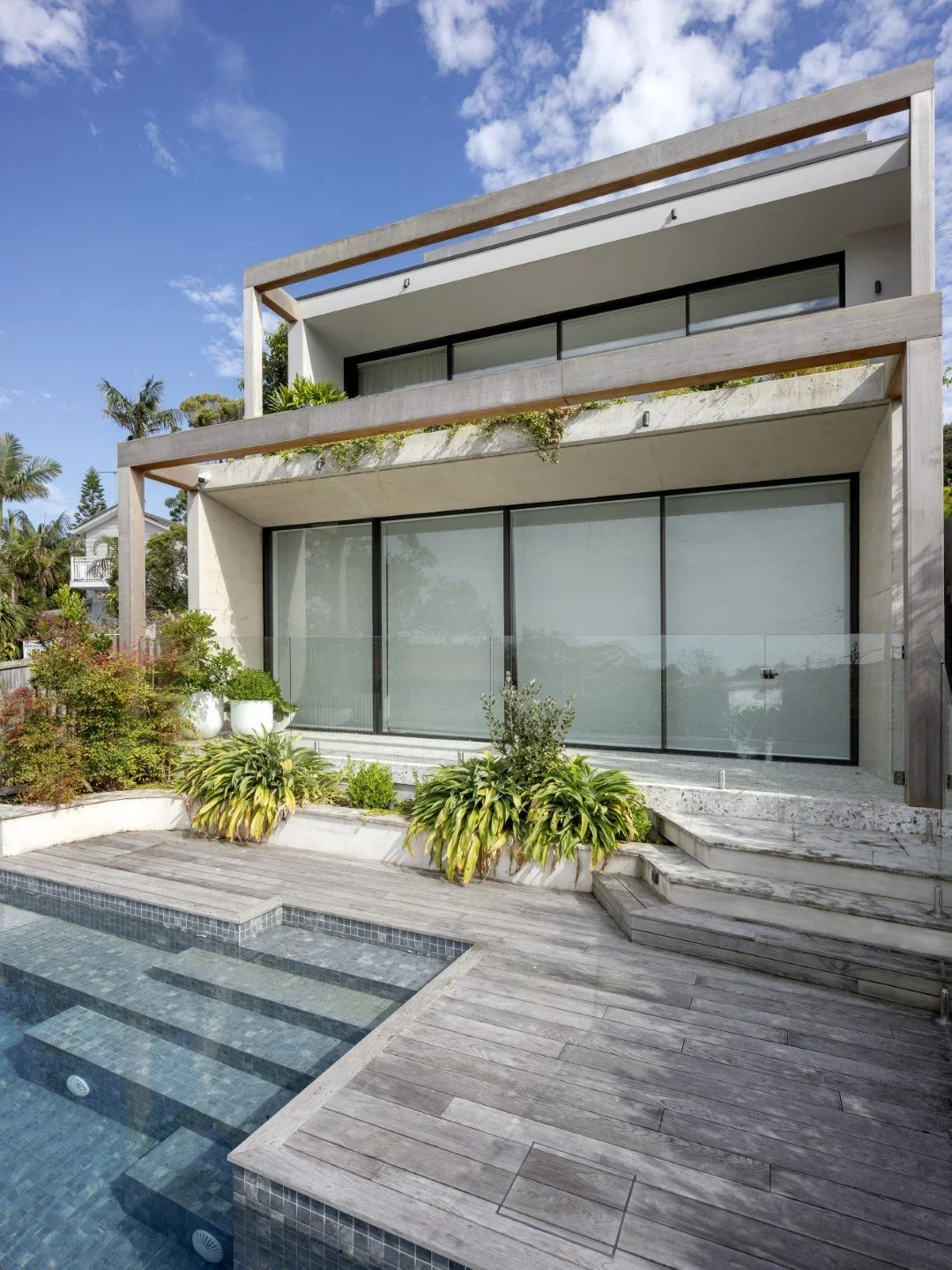 Modern house with large glass sliding doors and balconies, surrounded by plants and an outdoor pool with wooden decking.