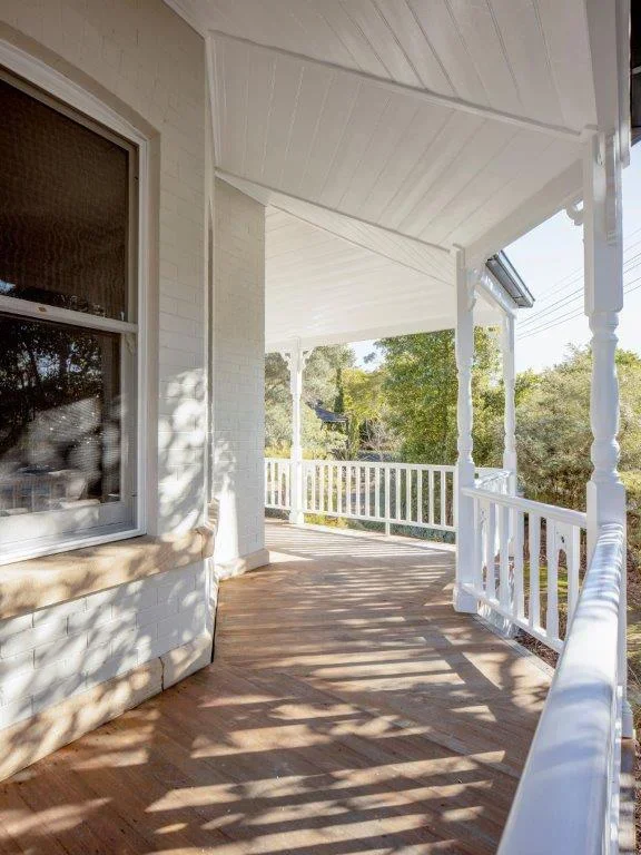 Front porch of a house with white railing and columns, wooden floor, and a window on the left side, with shadows of trees cast on the porch floor.