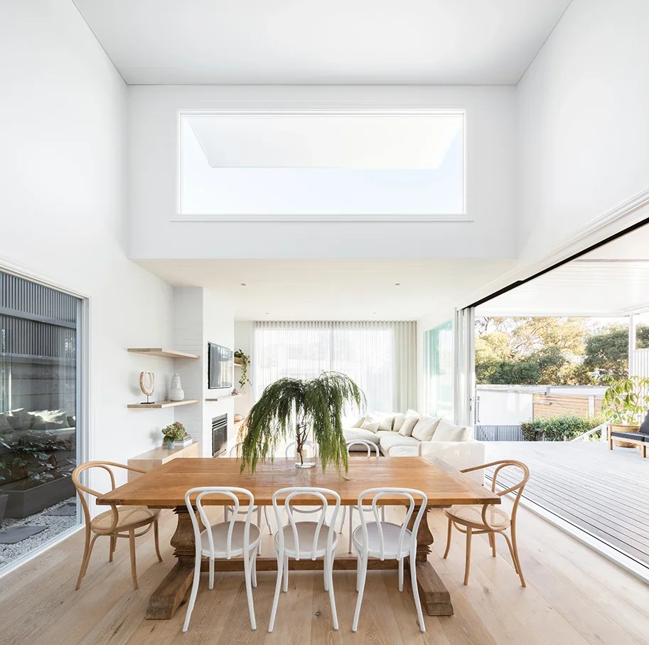 Bright, modern dining and living room with large sliding glass doors, a wooden dining table with eight chairs, a white sectional sofa, a fireplace, and a skylight.