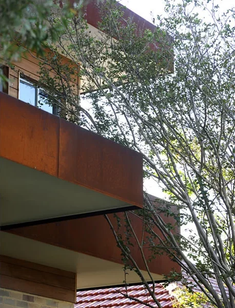 The corner of a modern building with large windows and a rust-colored metal balcony, partially obscured by a tree with green leaves.
