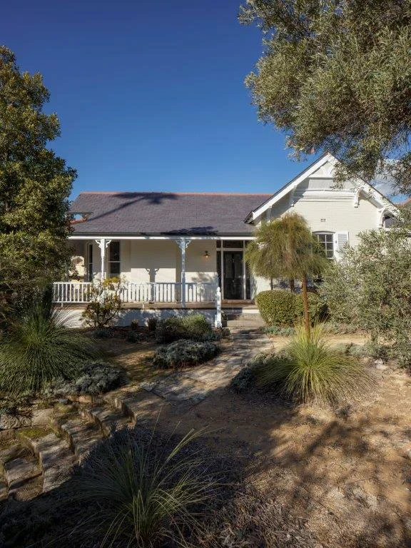 White house with porch and garden, trees, plants, blue sky.