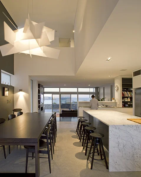 Modern kitchen and dining area with high ceilings, a large window view, white marble countertops, black stools, and a person sitting at the counter.