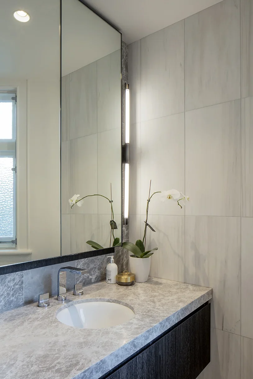 A modern bathroom vanity with a marble countertop, a sink, and decorative plants, with a large mirror and updated lighting.