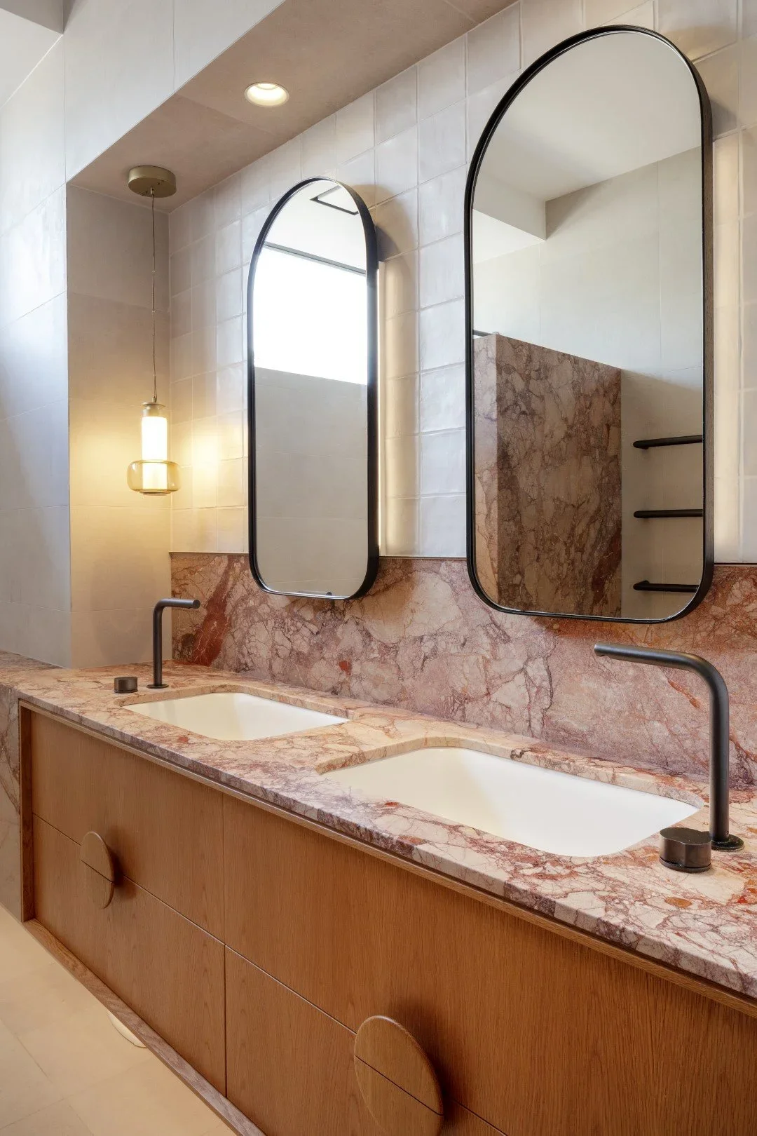 A modern bathroom vanity with a pink marble countertop and two rectangular white sinks. Two tall, narrow mirrors with black frames are mounted above the sinks, and a wooden cabinet with oval handles is below. A hanging pendant light and recessed ceil