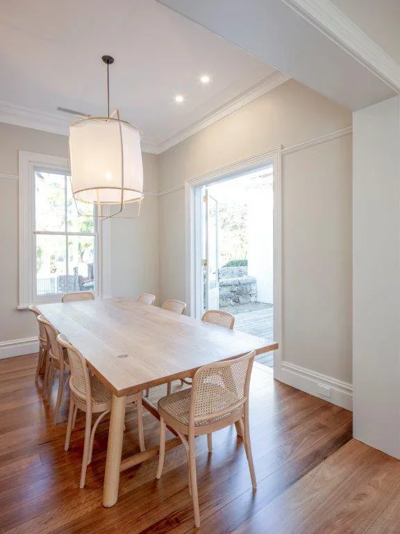 Dining room with a wooden table and eight chairs, large window and glass door, hardwood floor, white walls, pendant light, and outdoor patio visible through door.