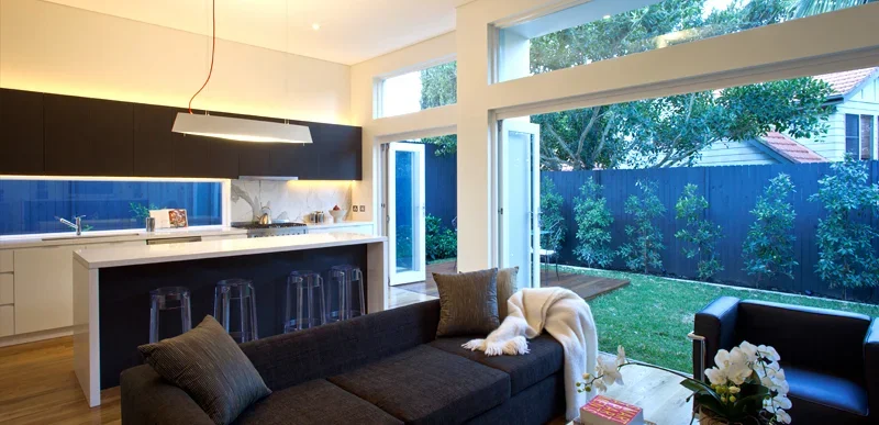 Open-concept living room and kitchen with large windows looking out onto a backyard with trees, a black couch with pillows and a blanket, a coffee table with flowers, and a modern kitchen with a white island and black cabinets.