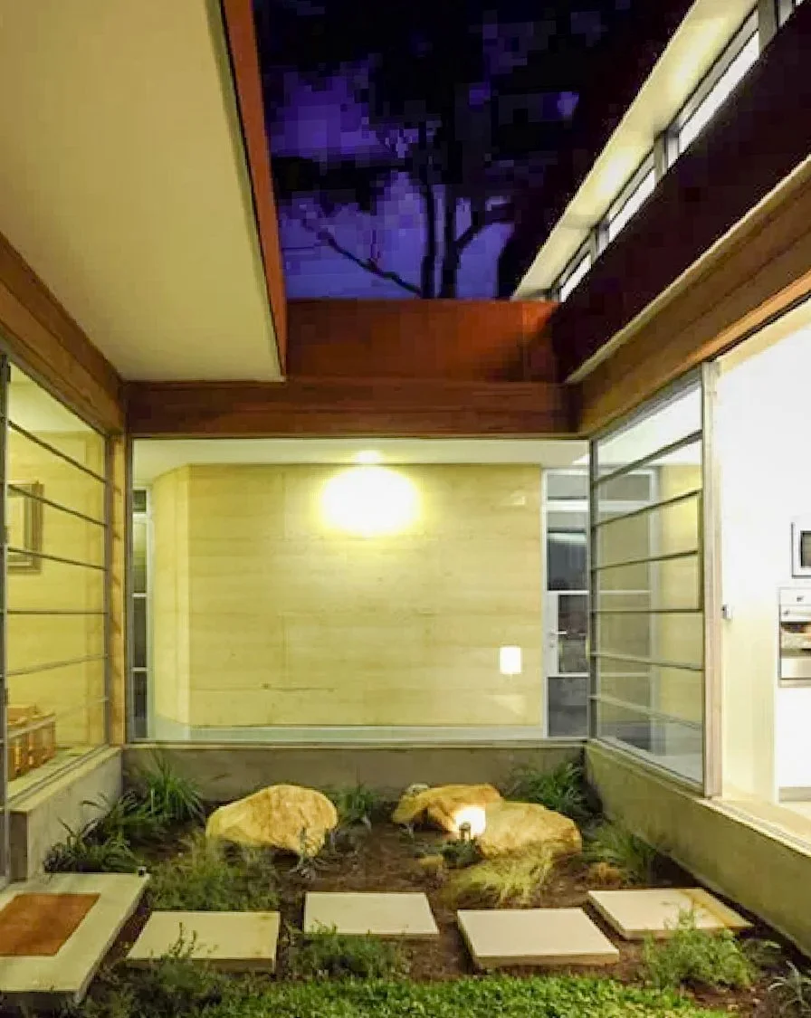 Interior view of a modern house with a small outdoor garden area featuring rocks and plants, visible through large glass windows at night.