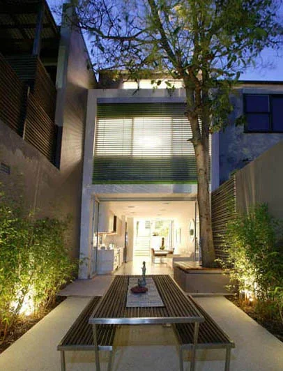Modern house with a patio, outdoor seating, tall trees, and illuminated landscaping at dusk.
