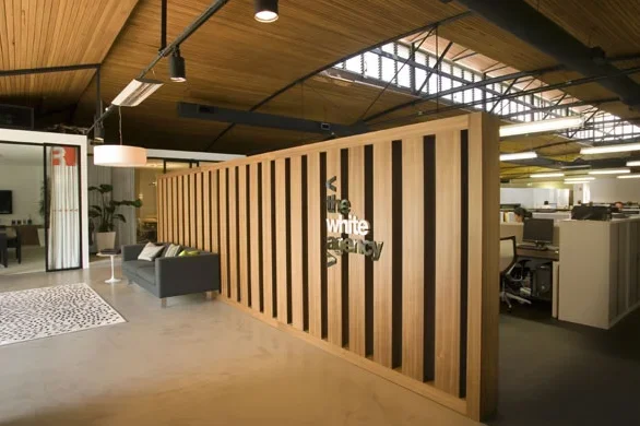 Office interior with a wooden slat wall displaying the text 'the white agency', a gray couch, a white rug, and workstations in the background.