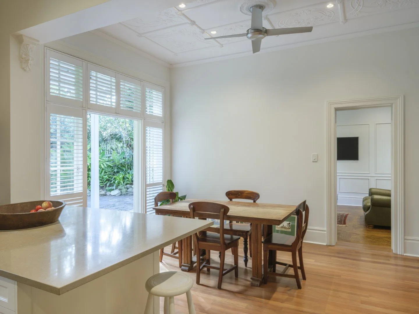Dining area with wooden table and chairs, large window with shutters, view of outdoor patio with plants, ceiling fan, adjacent living room with wall-mounted TV, hardwood flooring.