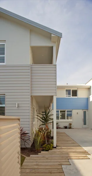 Exterior view of modern multi-story residential buildings with a wooden walkway, plants, and a cloudy sky.
