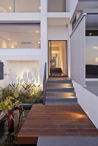 Exterior view of a modern house with stairs leading to a hallway, with plants and water feature nearby, and large windows giving a view inside.